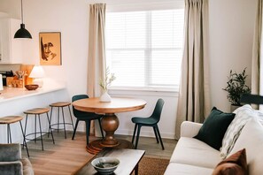 Ett modernt vardagsrum med ett runt matbord i trä, gröna stolar, en vit soffa och en köksdel med en hängande taklampa och en inramad tavla på väggen.
