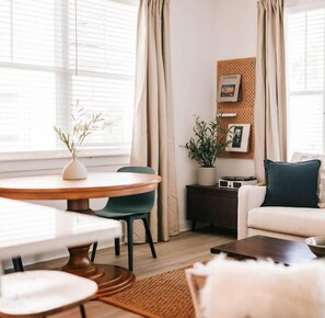 Uma sala de estar moderna com uma mesa de jantar redonda, um sofá e uma mesa de centro. Há uma planta em uma mesinha lateral e um porta-retrato na parede.