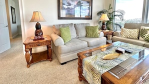 Ein Wohnzimmer mit einem beige farbenen Sofa, zwei Beistelltischen mit Lampen, einem Couchtisch mit tropischem Blattmuster und einem gerahmten Bild an der Wand.