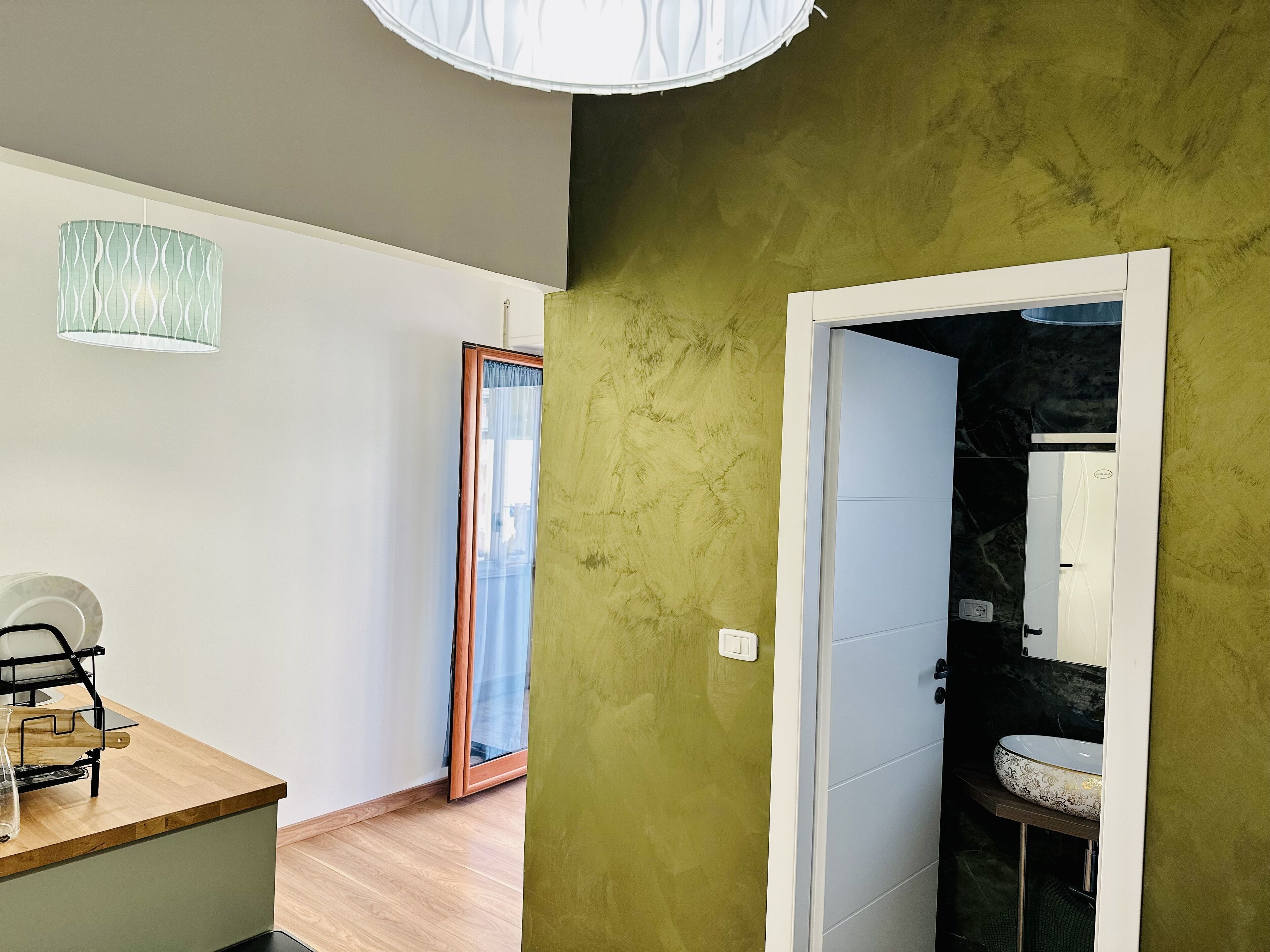 A room with a wooden desk, a white chair, a green accent wall, a bathroom with a sink and mirror, and a large window with a curtain.