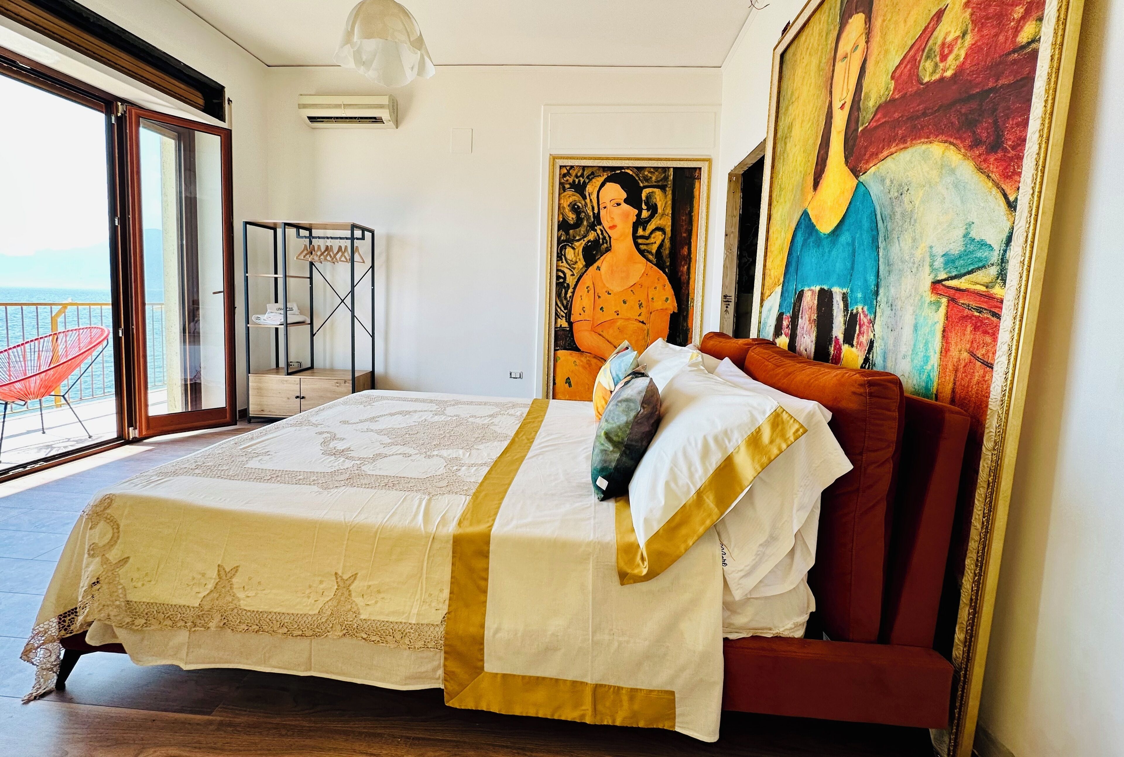 A bedroom with a large bed, a balcony with a red chair, and a painting on the wall.