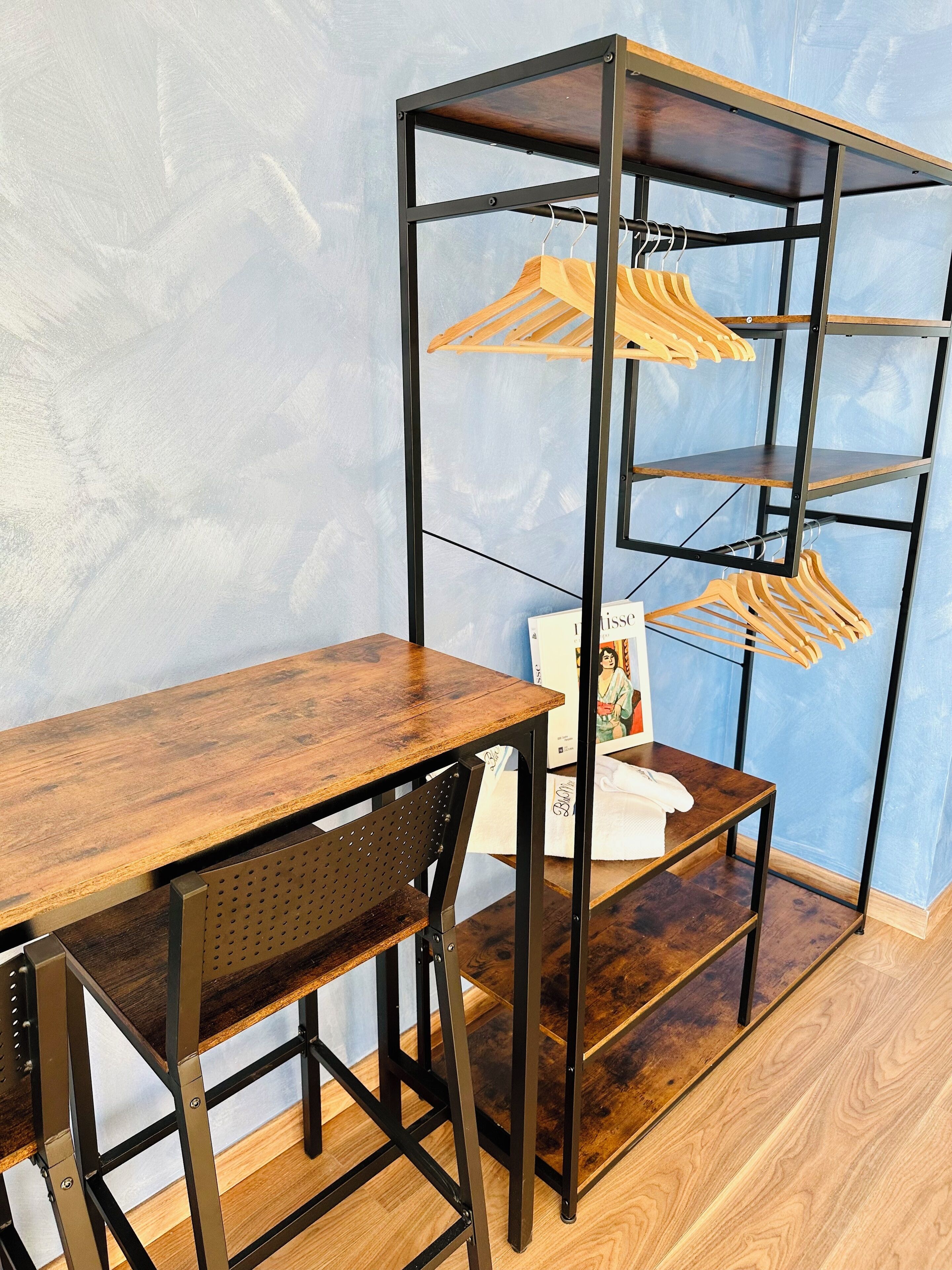 Quarto compacto com escrivaninha e cadeira de madeira, prateleira de metal com cabides de madeira e uma pequena mesa branca.