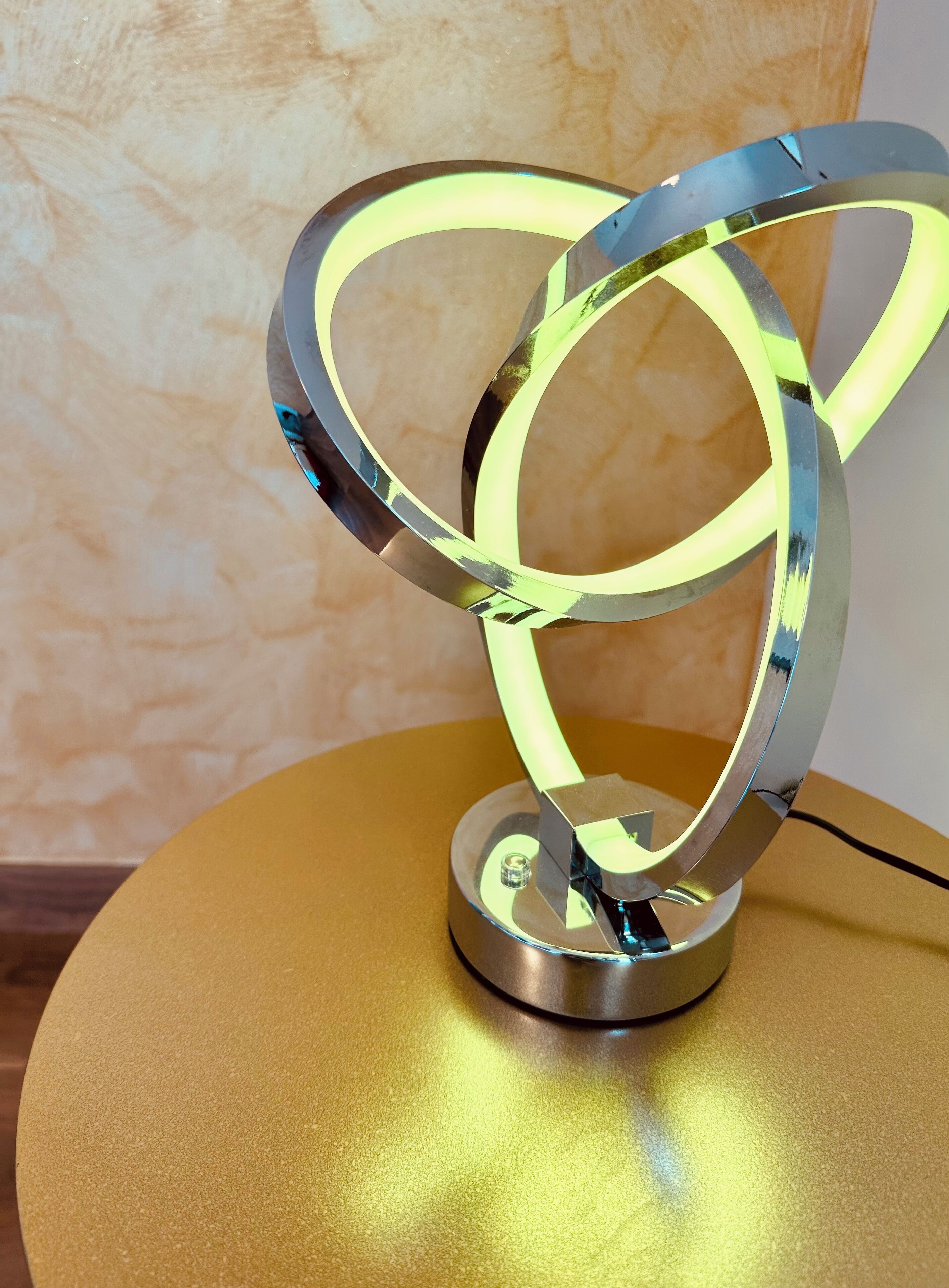 Uma luminária metálica circular com superfície refletora e elemento luminoso, fixada sobre uma mesa redonda dourada, em frente a uma parede texturizada.