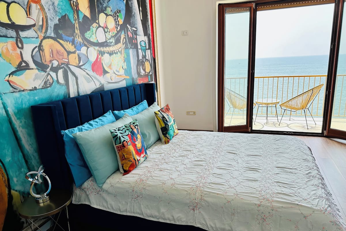 A bedroom with a bed, colorful pillows, a large window with a view of the sea, and a balcony with chairs.