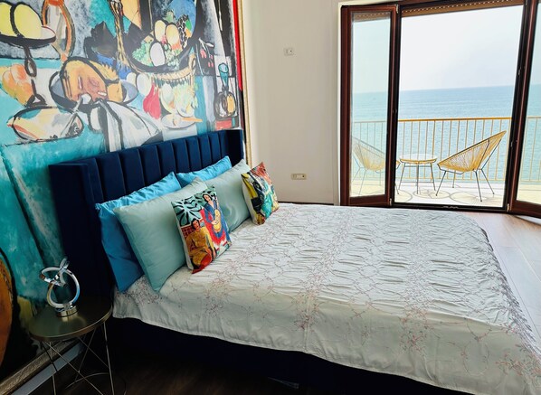 A bedroom with a bed, colorful pillows, a large window with a view of the sea, and a balcony with chairs. - BluMare ArtHouse (Salerno)