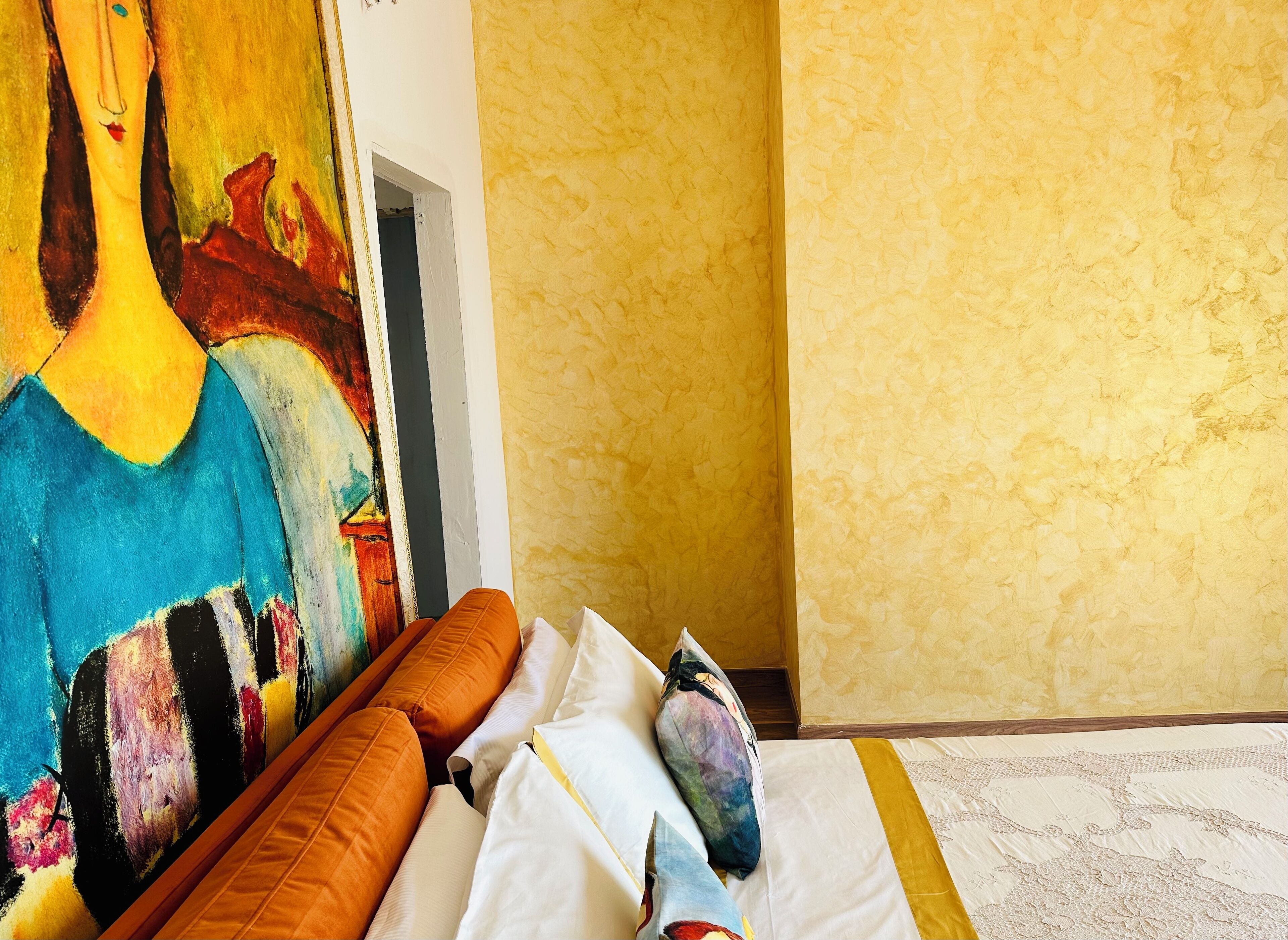 A hotel room with a large abstract painting on the wall, a bed with white linens and orange pillows, and a patterned rug.