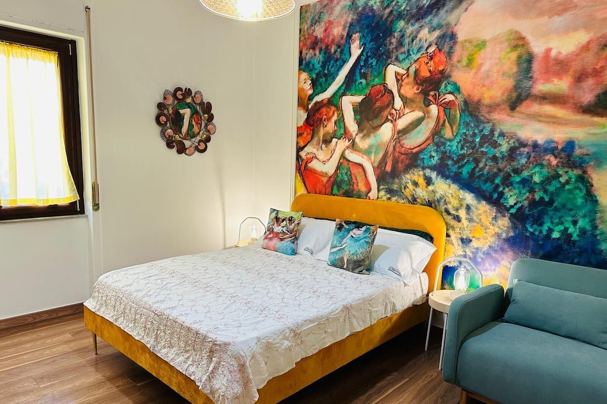 A bedroom with a bed, a blue armchair, a painting, and a wall clock.