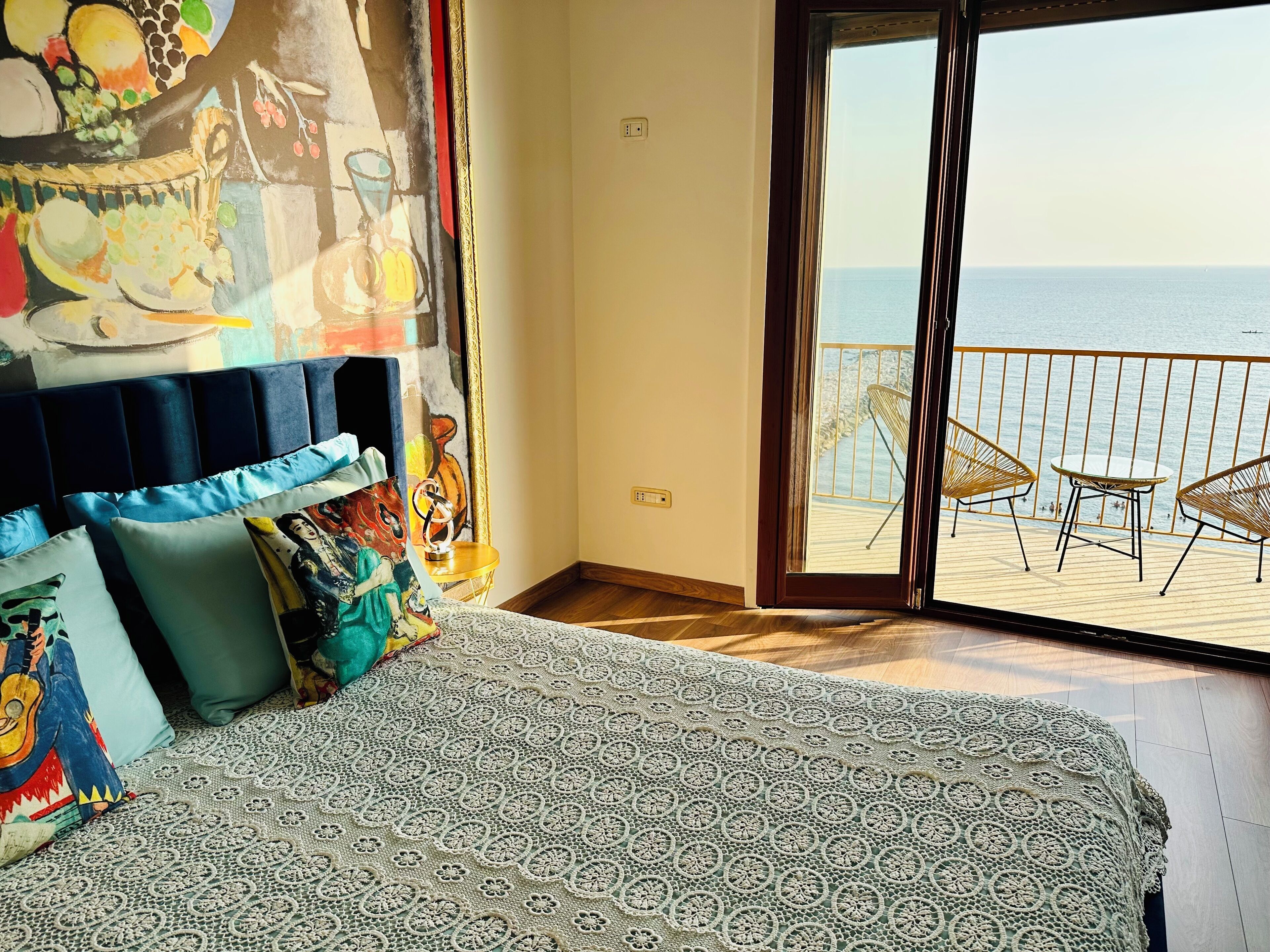 A bedroom with a bed, colorful pillows, a lace-patterned blanket, and a view of the sea through a sliding glass door.
