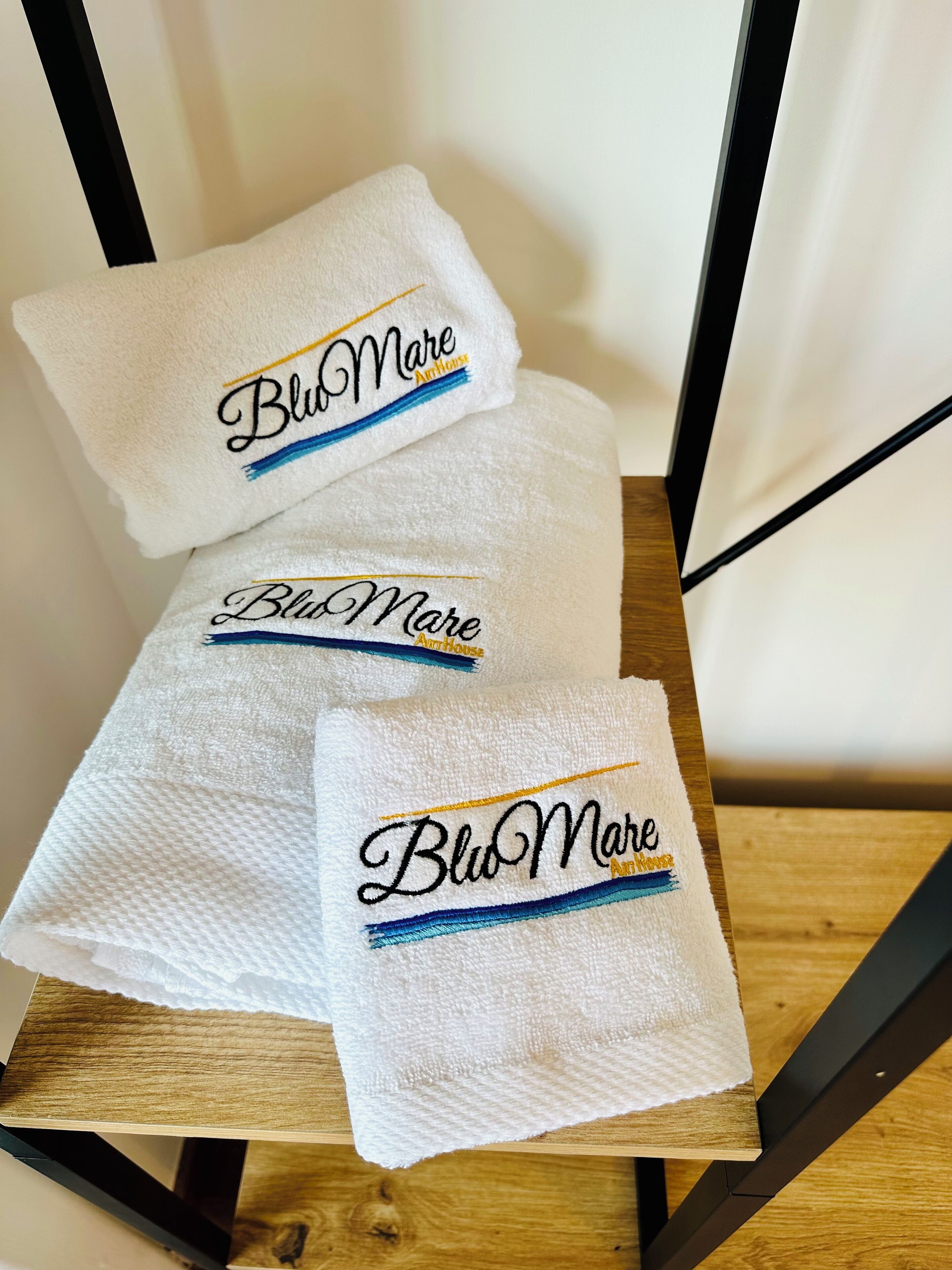Three white towels with embroidered text "Blu Mare" on a wooden chair.