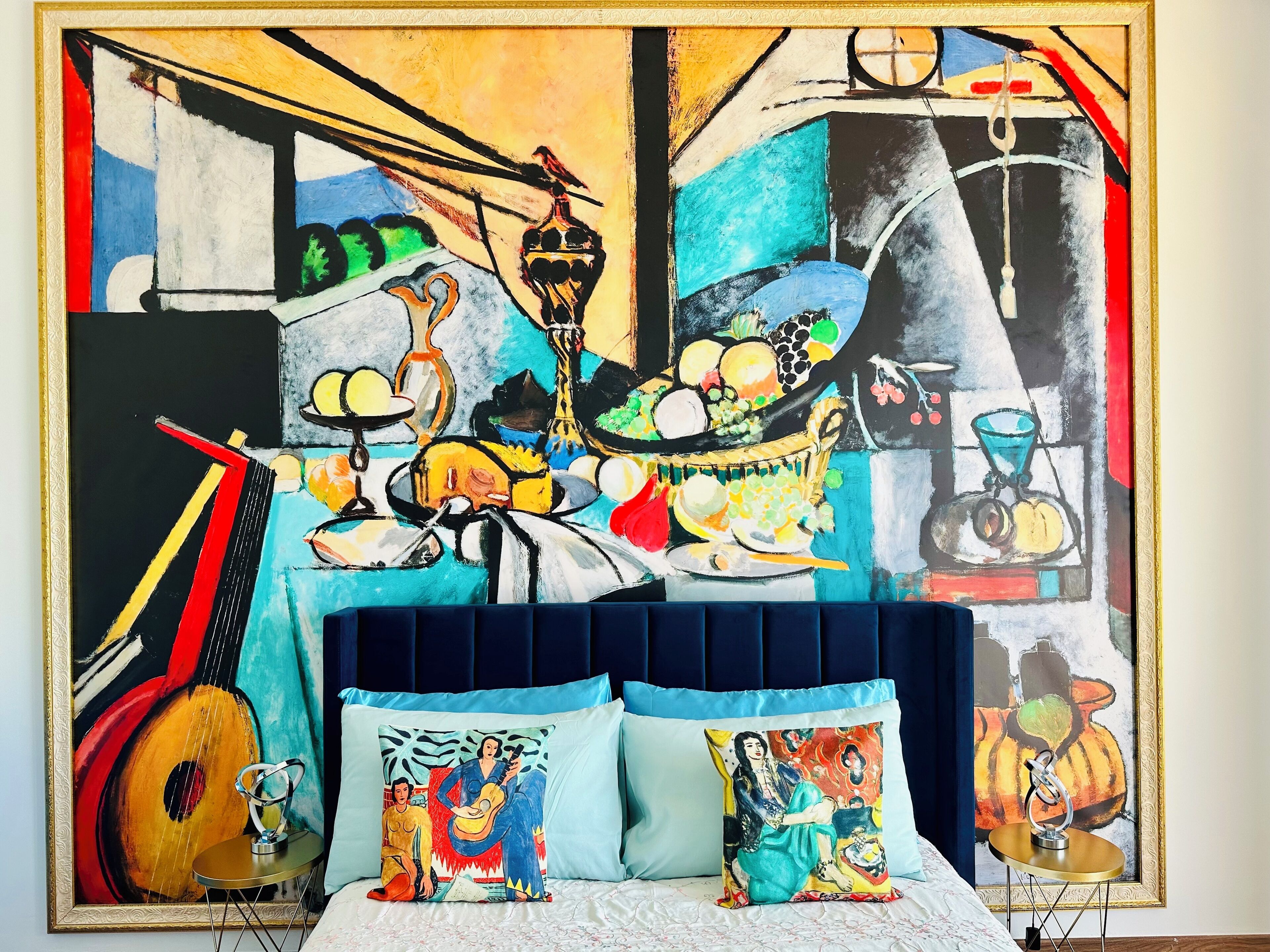 A bedroom with a large abstract painting on the wall, a bed with a blue headboard, and two pillows with colorful artwork.