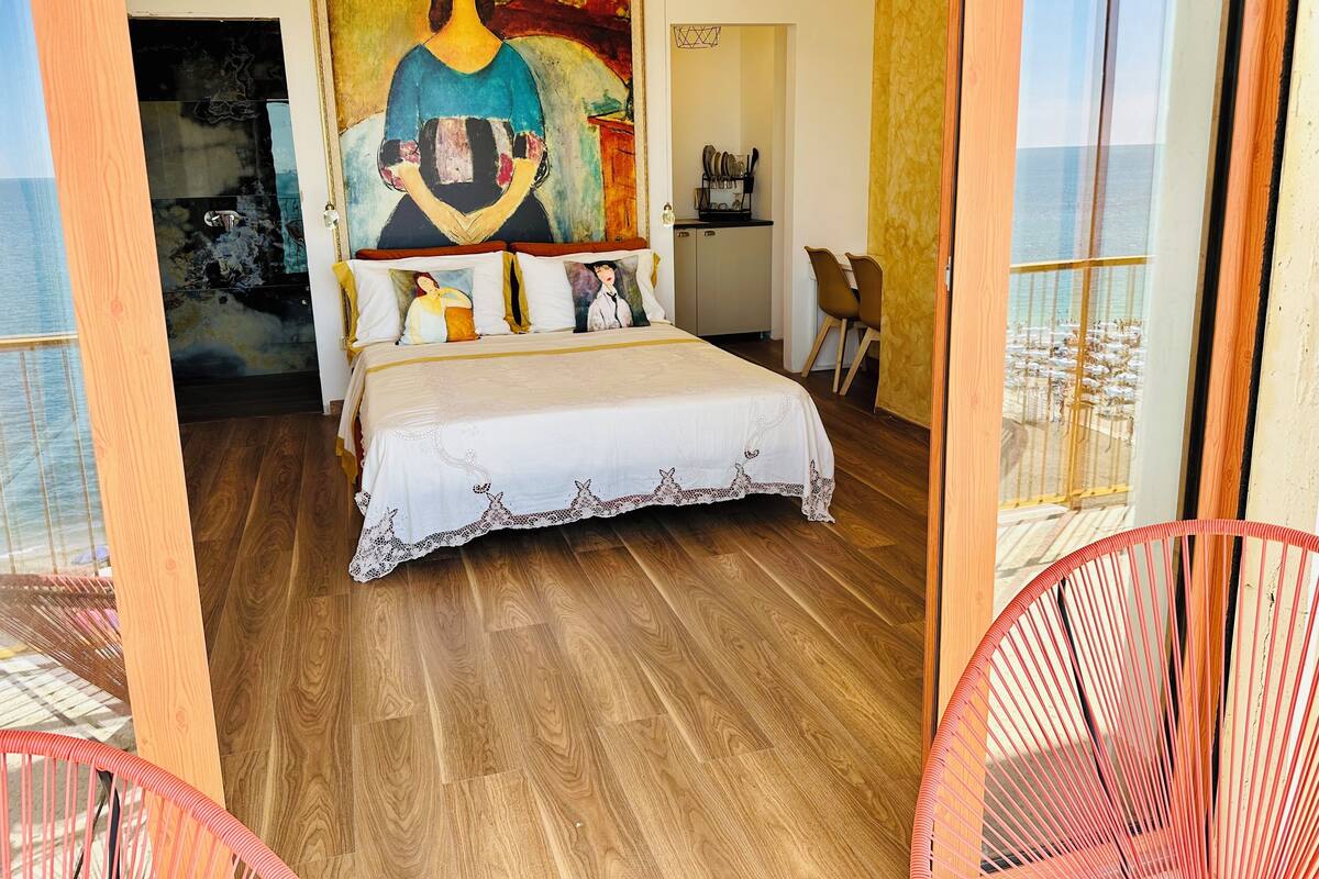 A bedroom with a large bed, a painting on the wall, and a view of the sea through a window.