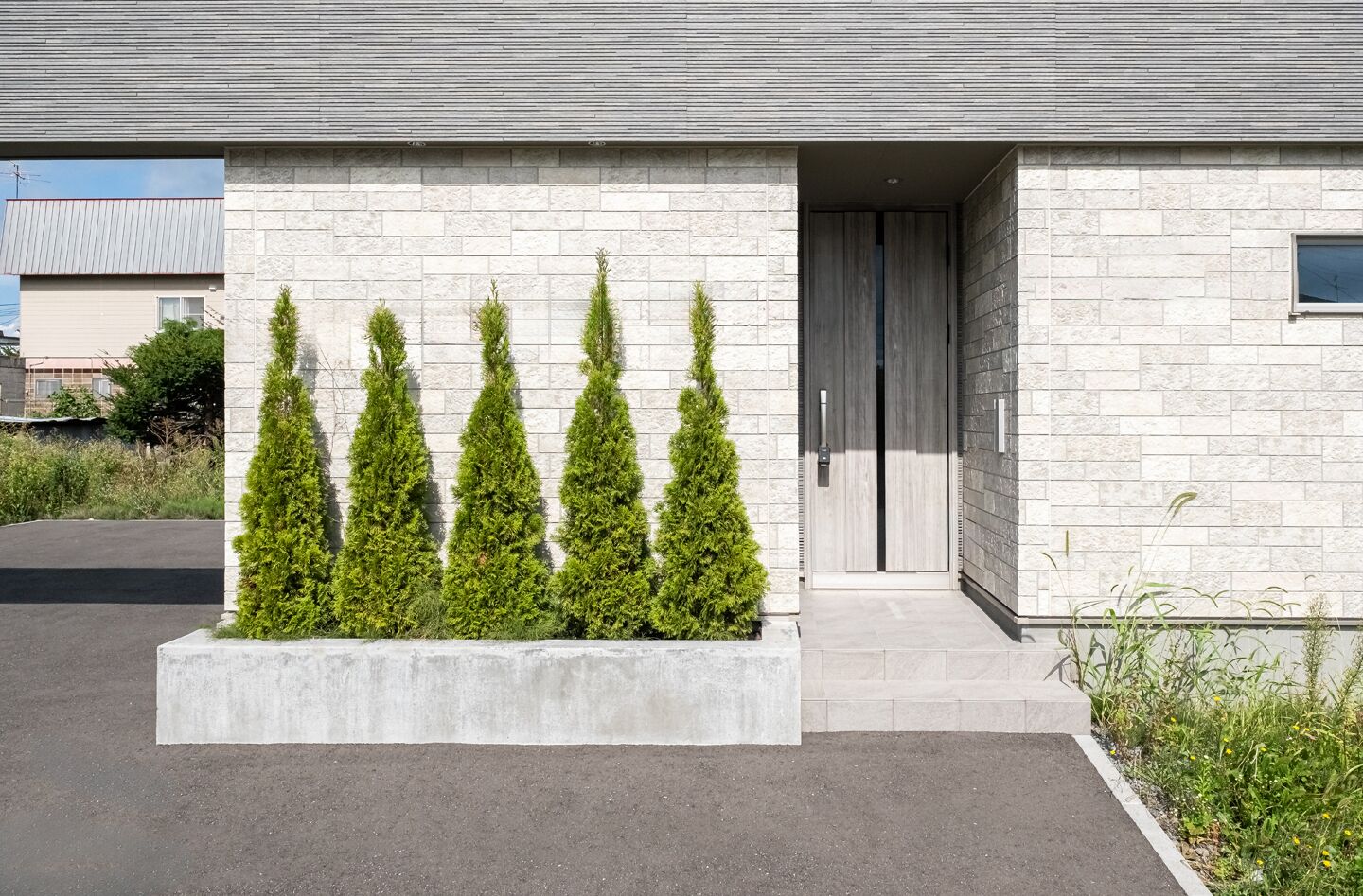Un edificio moderno con fachada de piedra, una jardinera de hormigón con árboles de hoja perenne y una puerta de madera.