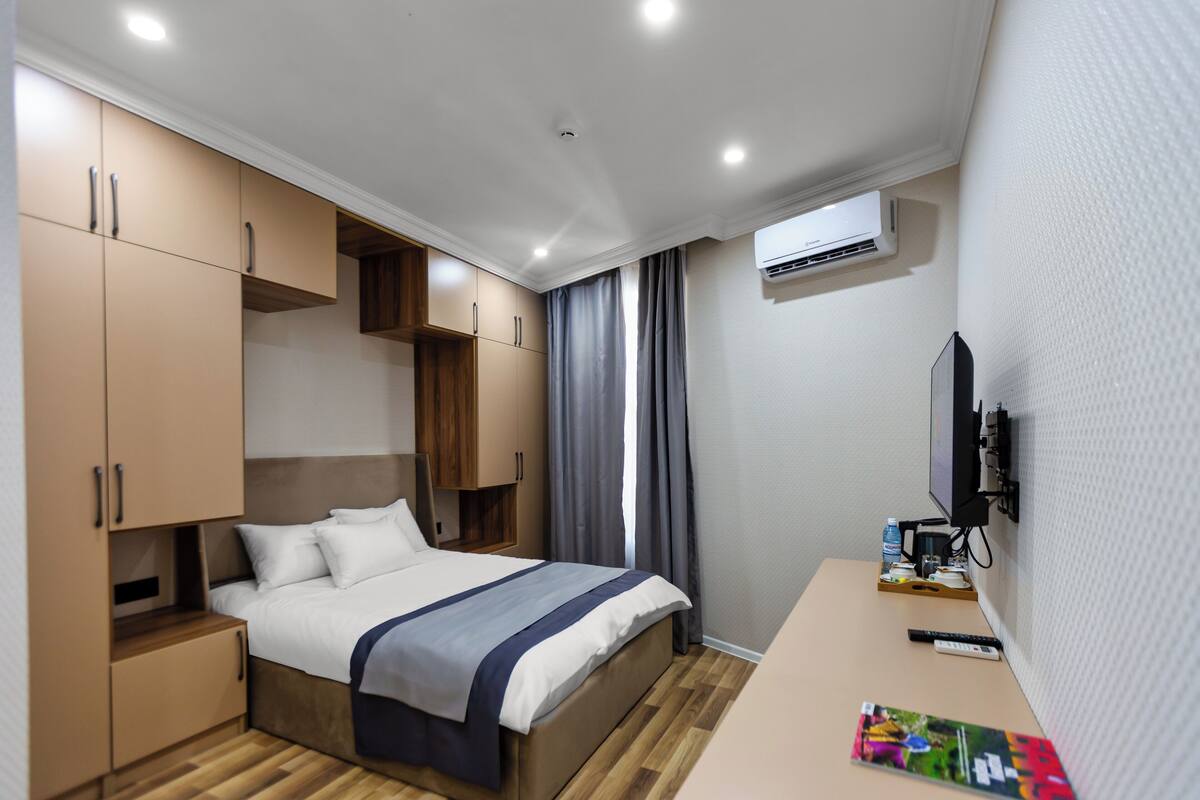 Un dormitorio con una cama, un armario de madera, un escritorio con un control remoto, una revista y un equipo de aire acondicionado.