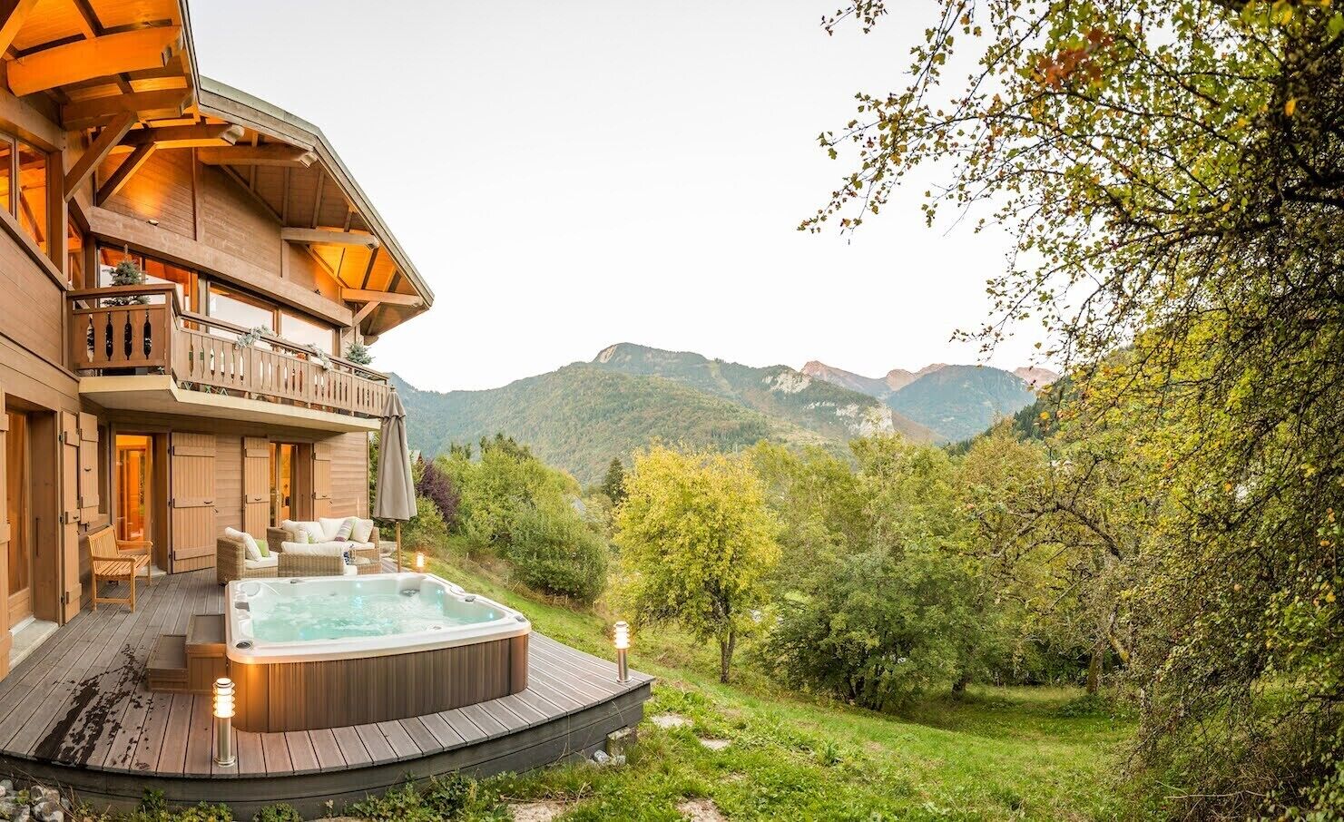 A wooden building with a hot tub and outdoor seating, surrounded by greenery and mountains.