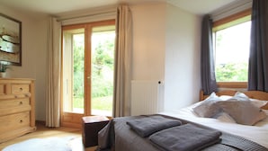 Ein Schlafzimmer mit einem Bett, einer Holzkommode, einem Fenster mit Vorhängen und Blick auf Grünfläche.