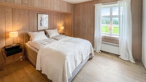 Ein Schlafzimmer mit einer Holzwand, einem großen Fenster mit weißen Vorhängen, einem Bett mit weißer Bettwäsche und zwei Nachttischlampen.