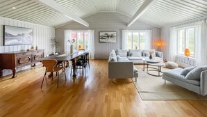 Ein geräumiger Wohnbereich mit Holzboden, einem Esstisch mit Stühlen, einer Couch und einem Couchtisch. Große Fenster mit Vorhängen und eine Decke mit sichtbaren Balken.