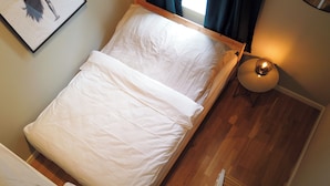 Una cama individual con ropa de cama blanca en una habitación con suelo de madera y una mesita de noche con lámpara.
