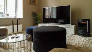Una sala de estar con un televisor de pantalla plana, un mueble blanco, un reposapiés negro, una mesa de centro de vidrio y un ventanal grande.