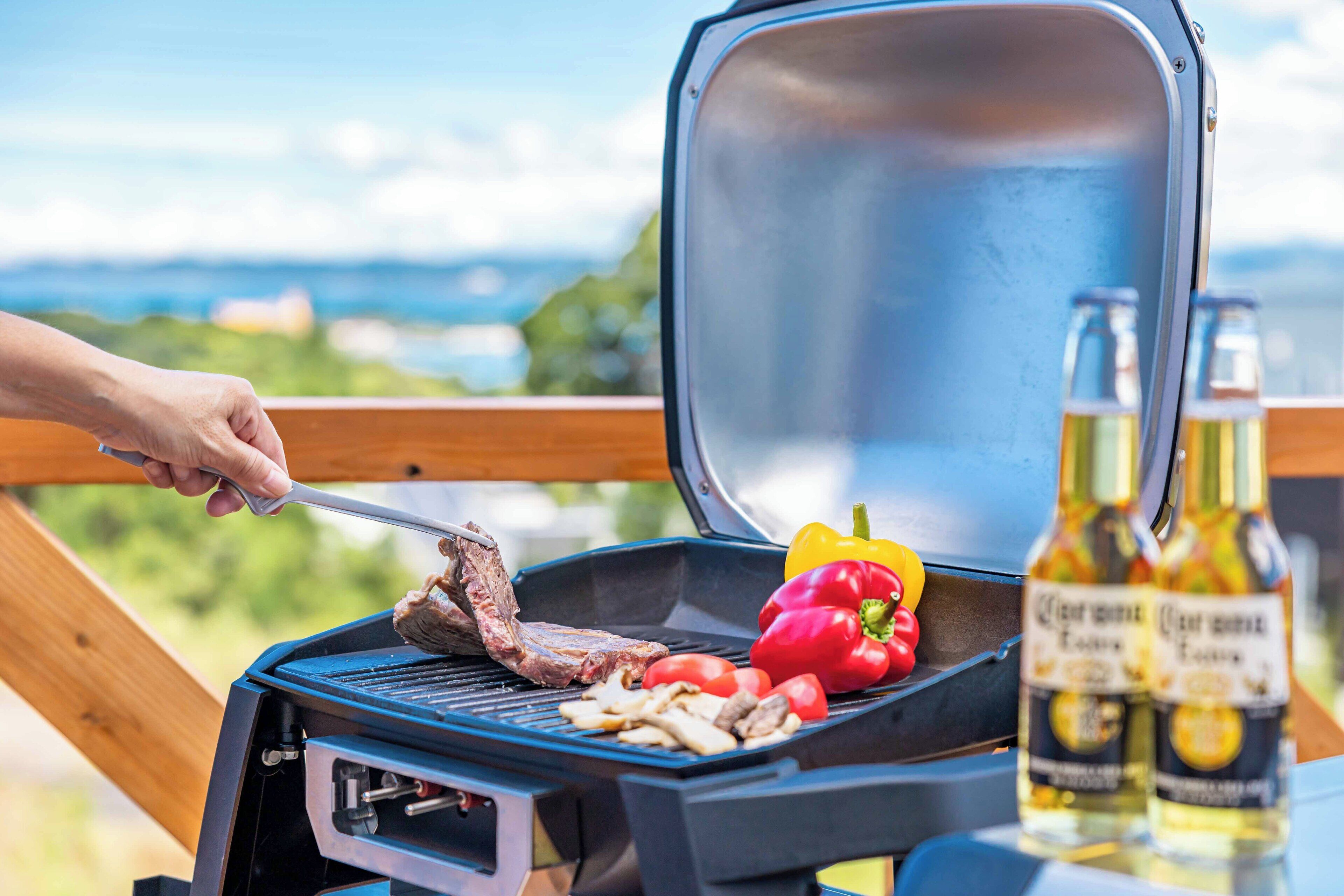 A grill with meat, mushrooms, and a red bell pepper, and two bottles of beer on a table with a wooden railing and a scenic view in the background.
