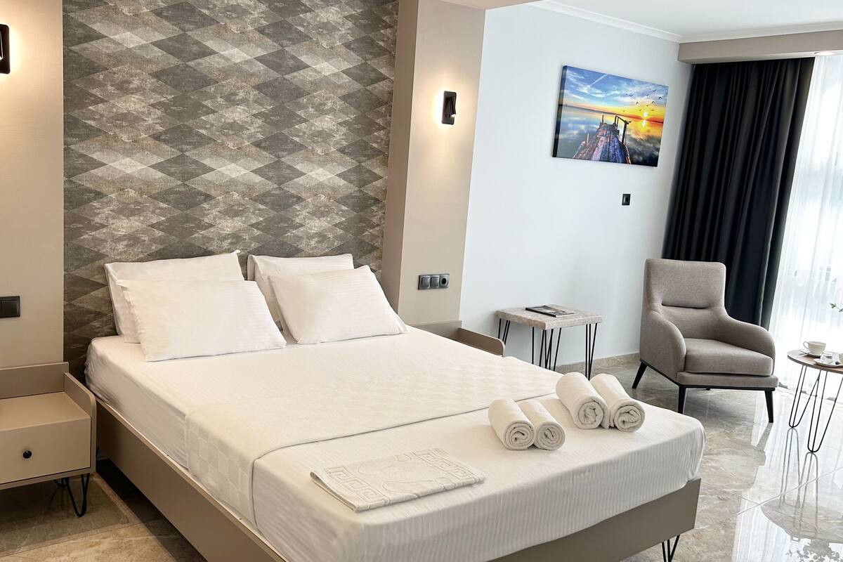 A modern hotel room with a large bed, a bedside table with a phone, a small side table, a chair, and a painting on the wall.
