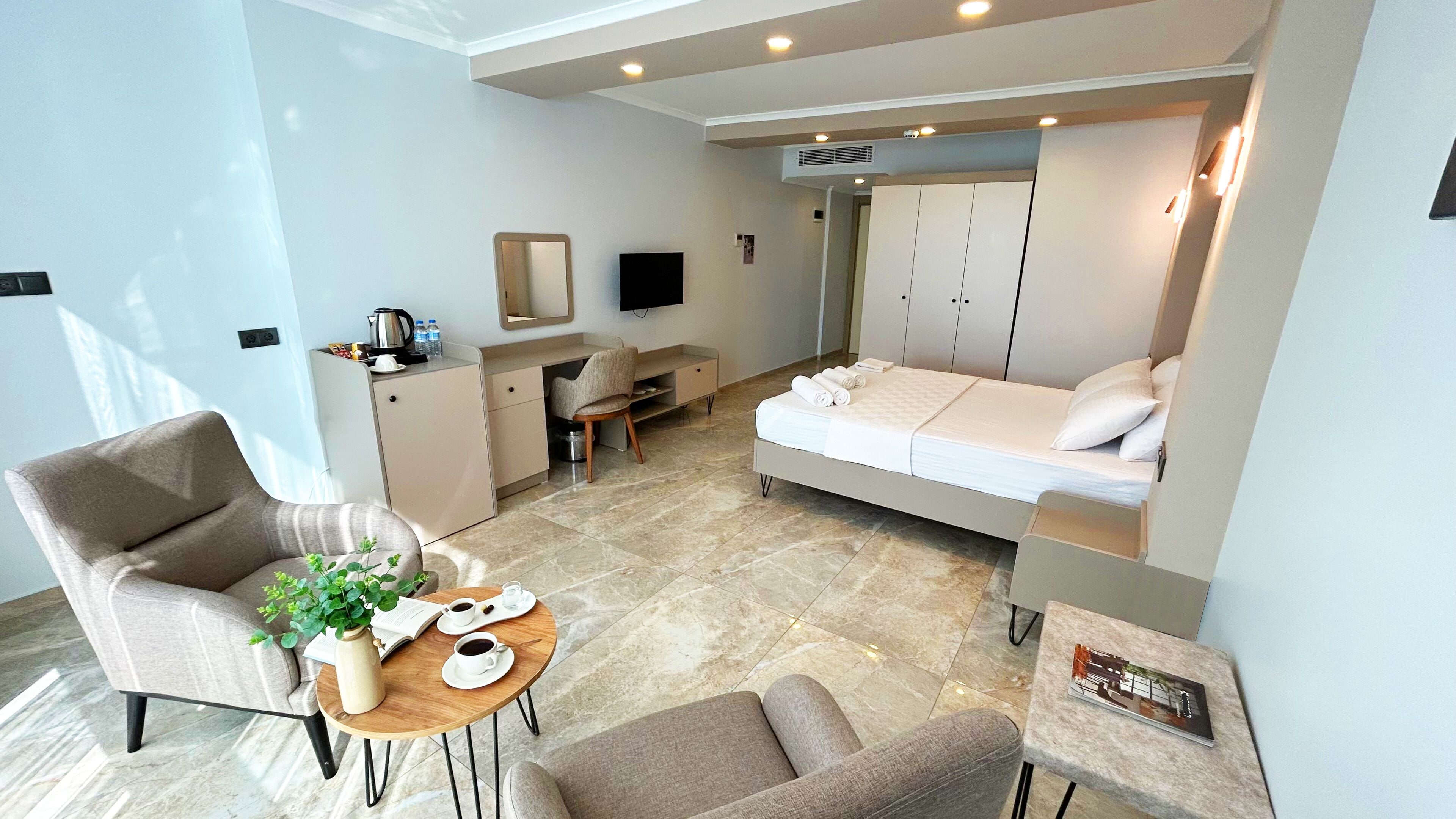 Une chambre d’hôtel de taille réduite, comprenant un lit, un bureau, une chaise, une petite table avec une plante et des tasses, ainsi qu’une armoire.