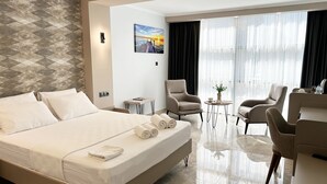 A hotel room with a large bed, two armchairs, a small table, and a view of the outdoors through sheer curtains.