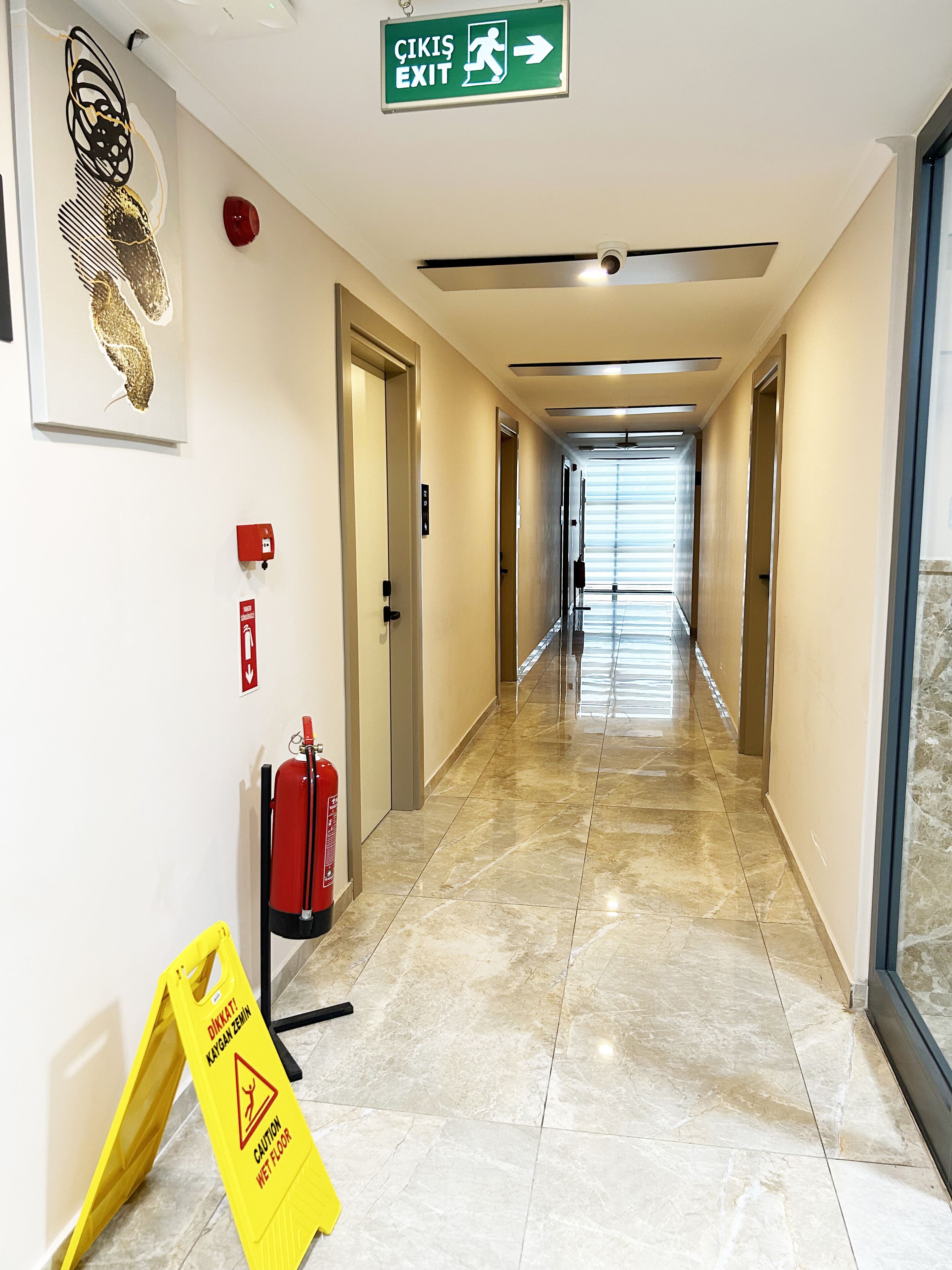 Un pasillo de hotel con un letrero de salida verde, extintor, señal de piso mojado y detector de incendios.