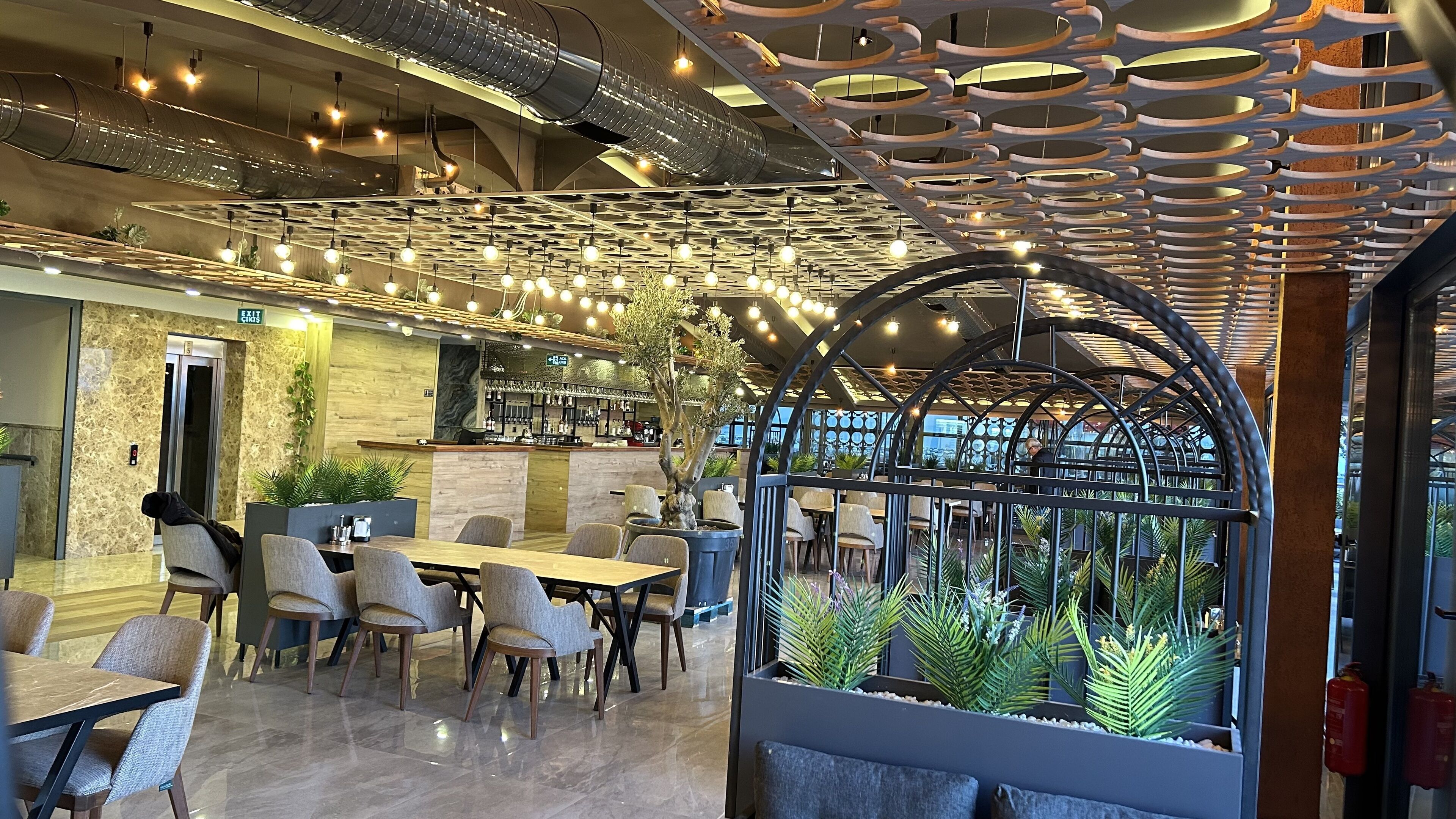 A modern restaurant with a bar area, seating with a view of indoor plants, and a unique ceiling design.