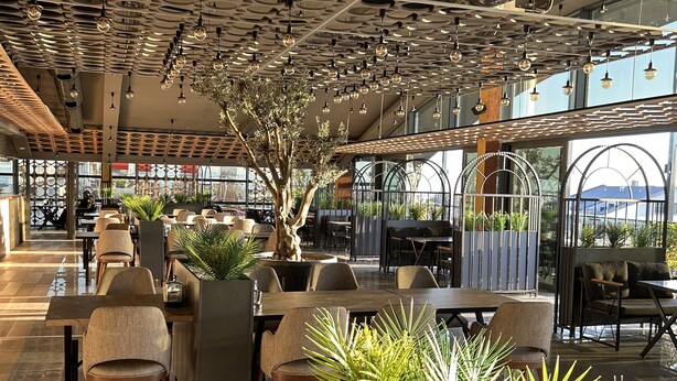 A modern restaurant with high ceilings, hanging spherical lights, and a central tree. Tables and chairs are arranged for dining.