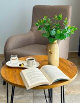 A cozy reading nook with a chair, a wooden table, a vase with green plants, a book, a cup of coffee, and a glass of water.