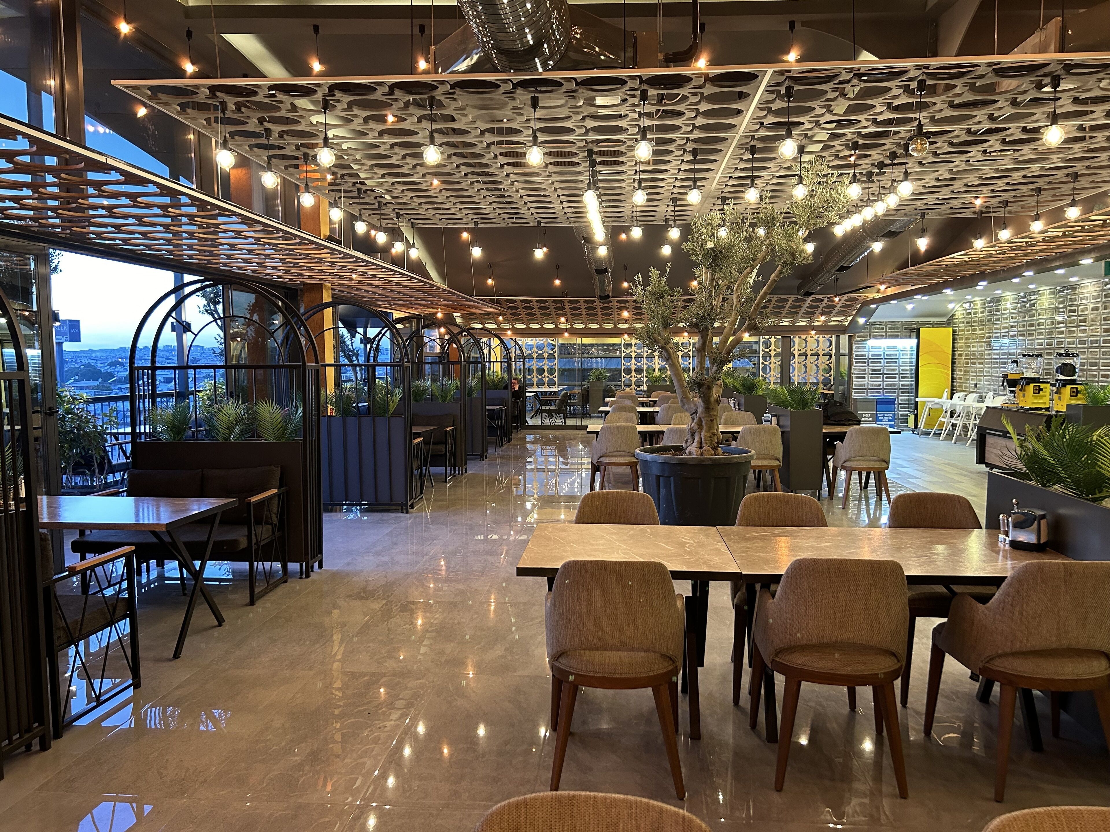 A modern restaurant with a high ceiling, pendant lighting, and a central tree planter. Tables and chairs are arranged for dining.