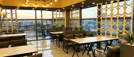 A modern indoor dining area with high ceilings, large windows, and a city view.