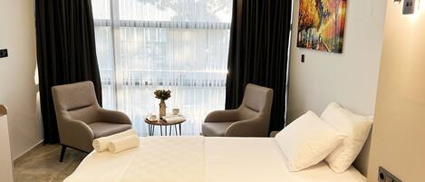 Une chambre d’hôtel avec un grand lit, deux fauteuils, une petite table avec un vase et une tasse, et un tableau au mur.