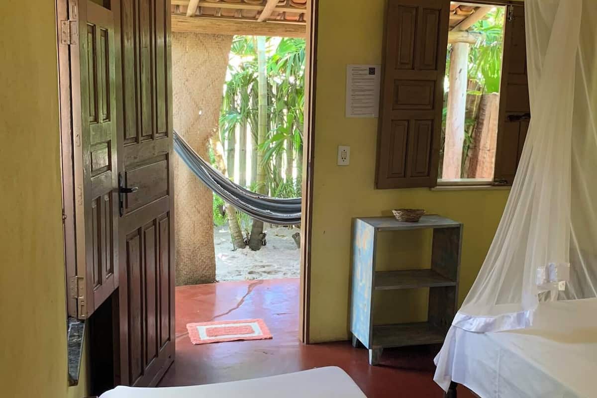 A room with a bed, a hammock, a small table, and a window with a view of greenery.
