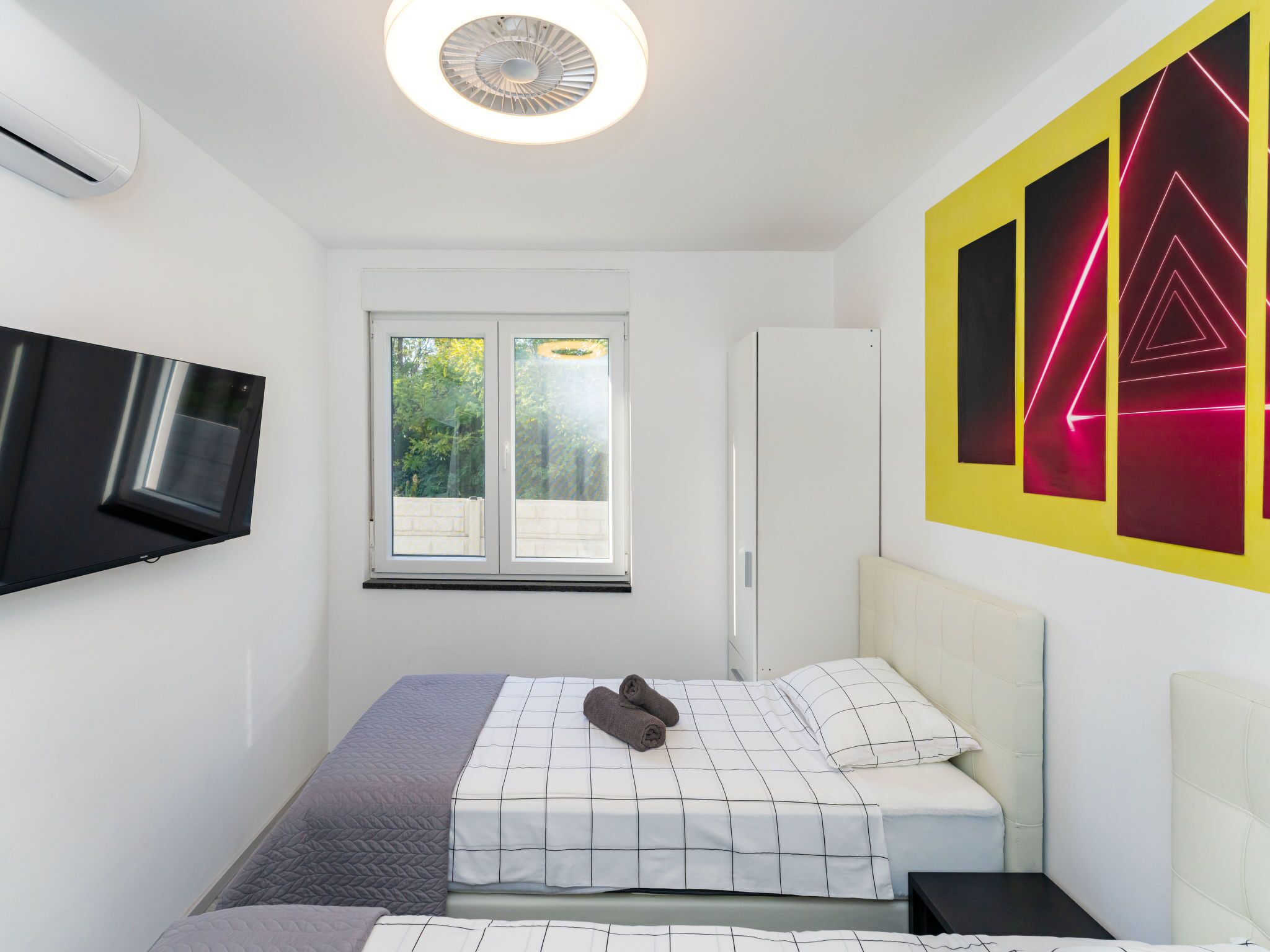 A modern bedroom with a bed, a TV mounted on the wall, and a window with a view of greenery.