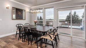 A dining area with a dark wooden table and chairs, a chandelier, and a large window with a view of a deck and trees. - Vineyard Vista Retreat | Close to Wineries, Lakes, & More! Sleeps 6, Hot Tub (Clements)