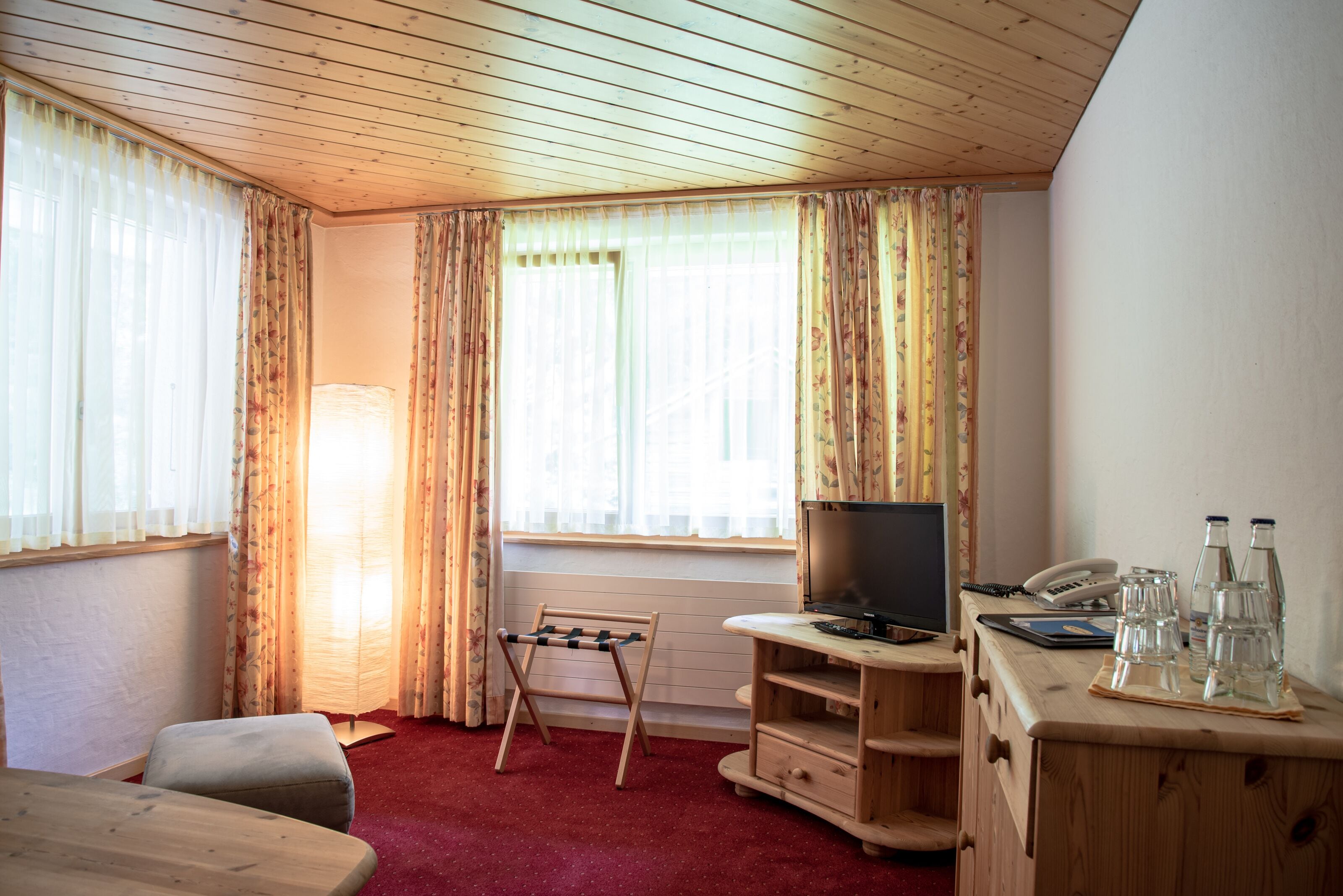 Een kamer met een houten plafond, een televisie op een houten dressoir, een kleine bank, een houten tafel met glazen en flessen, en een raam met gordijnen.