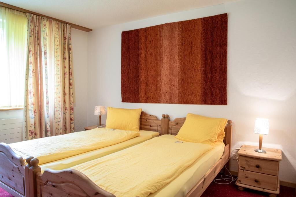 A room with two beds, each with a wooden headboard and yellow bedding. There is a wall-mounted artwork above the beds and a bedside table with a lamp on the right.