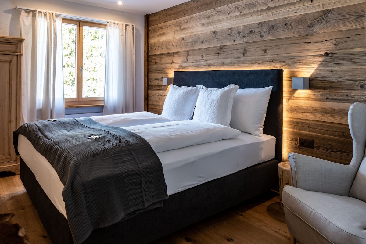 Ein ordentlich bezogenes Bett mit weißen und grauen Textilien, ein Holz-Kopfteil und ein Nachttisch mit Lampe.