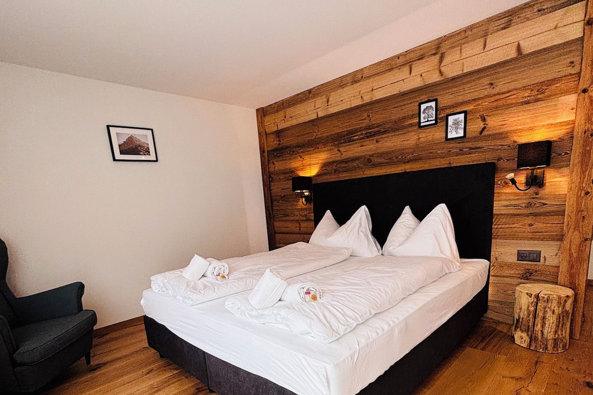 Ein Schlafzimmer mit einem hölzernen Kopfende, einem Bett mit weißen Leinen, einem dunklen Sessel und einem kleinen Holzschemel.