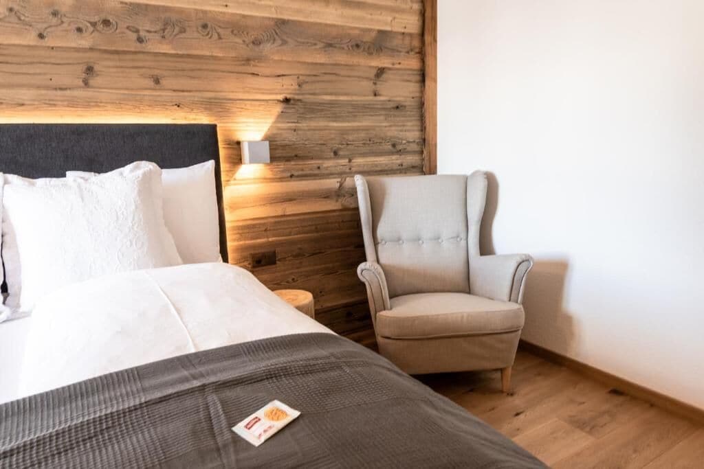 A bedroom with a wooden headboard, a bed with white bedding, a grey throw blanket, and a beige armchair.