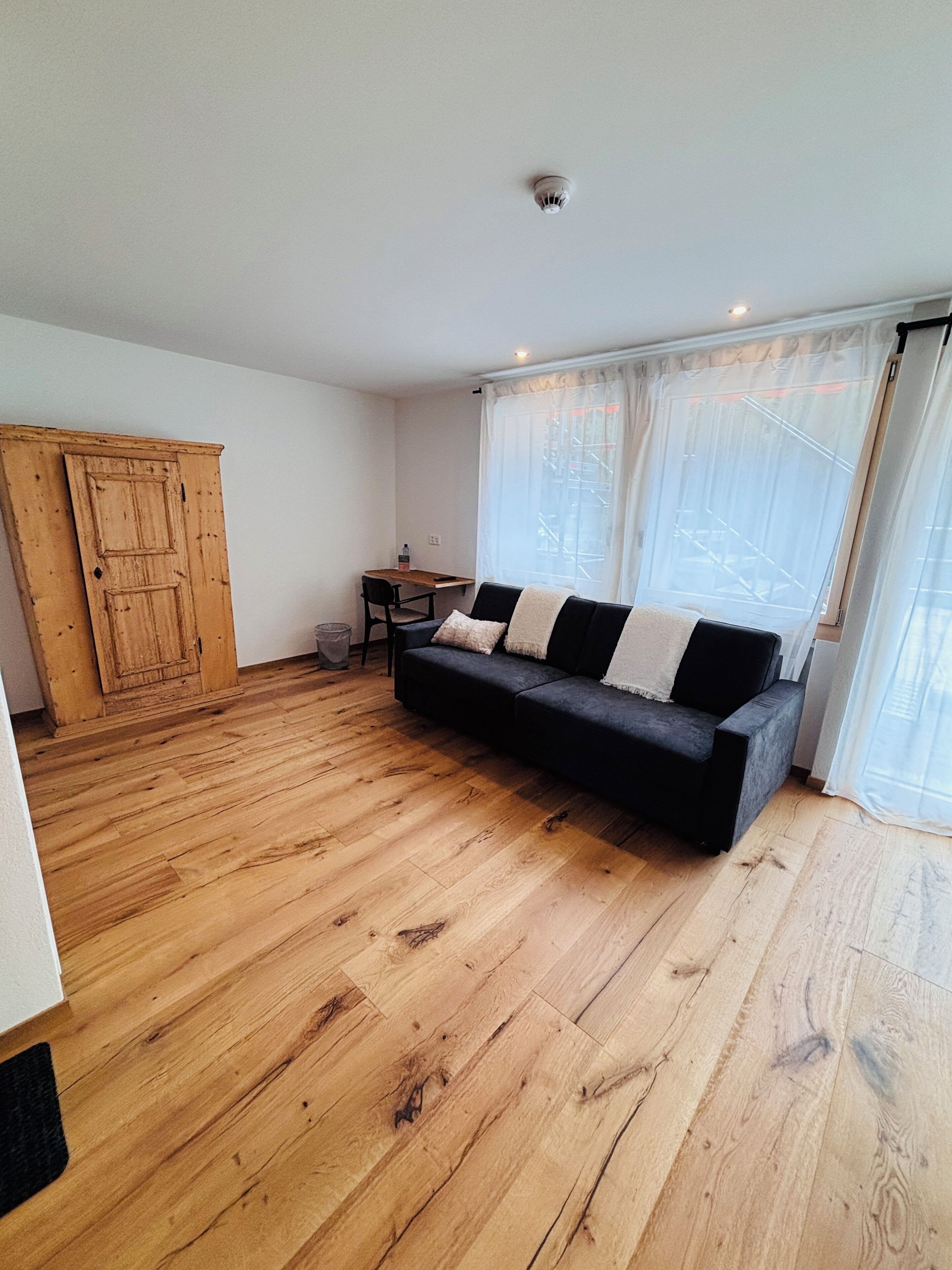 A spacious room with a wooden floor, a black sofa with cushions, a wooden wardrobe, a small table with a chair, and a window with sheer curtains.