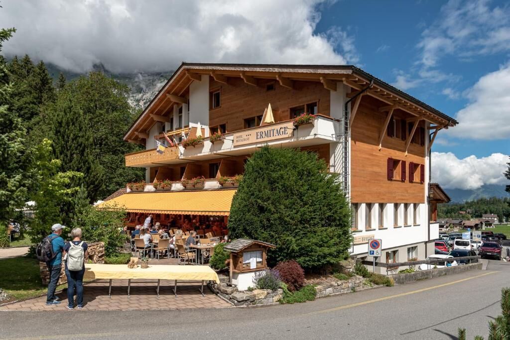 A wooden building with a restaurant named "ERMITAGE" on the ground floor, featuring outdoor seating and a mountain backdrop.