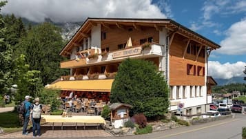 A wooden building with a restaurant named "ERMITAGE" on the ground floor, featuring outdoor seating and a mountain backdrop.