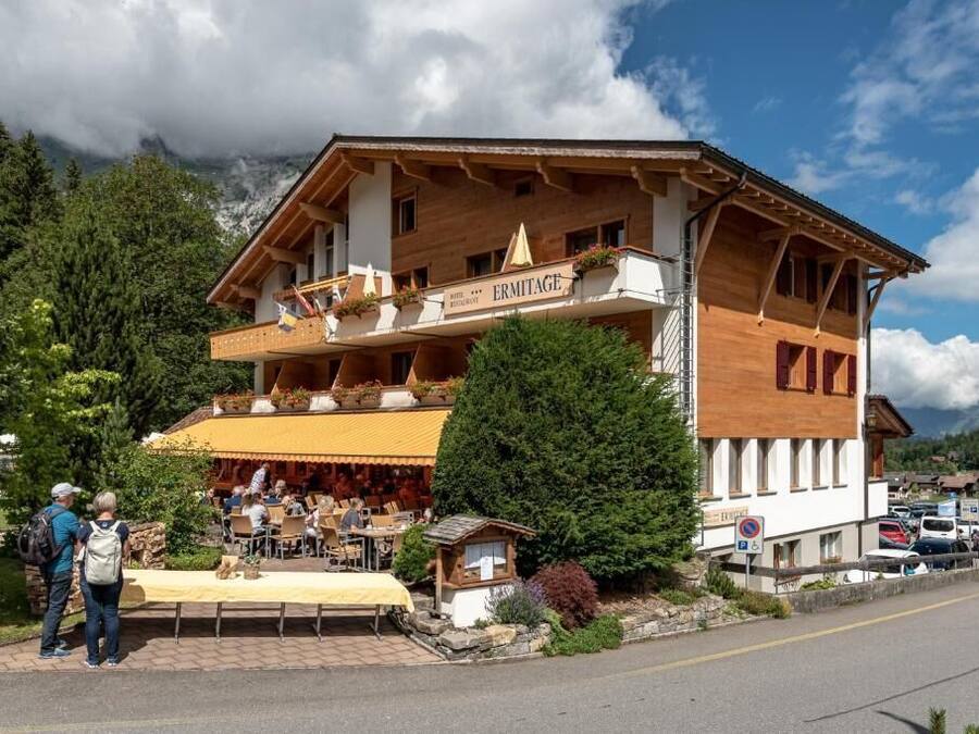 A wooden building with a restaurant named "ERMITAGE" on the ground floor, featuring outdoor seating and a mountain backdrop.