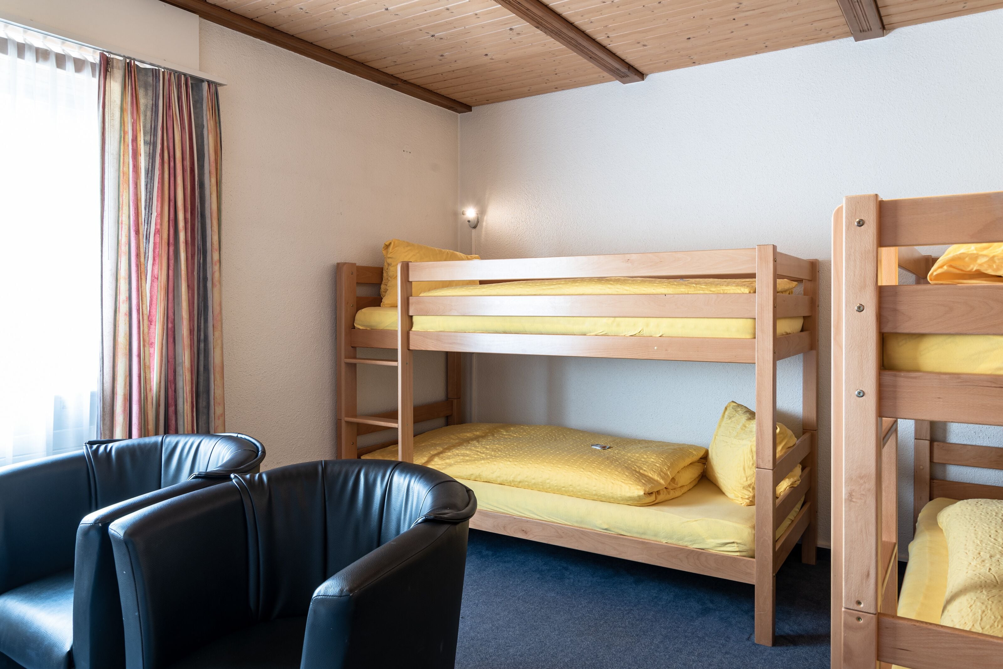 A bunk bed room with a wooden ceiling, a black leather chair, and a window with curtains.