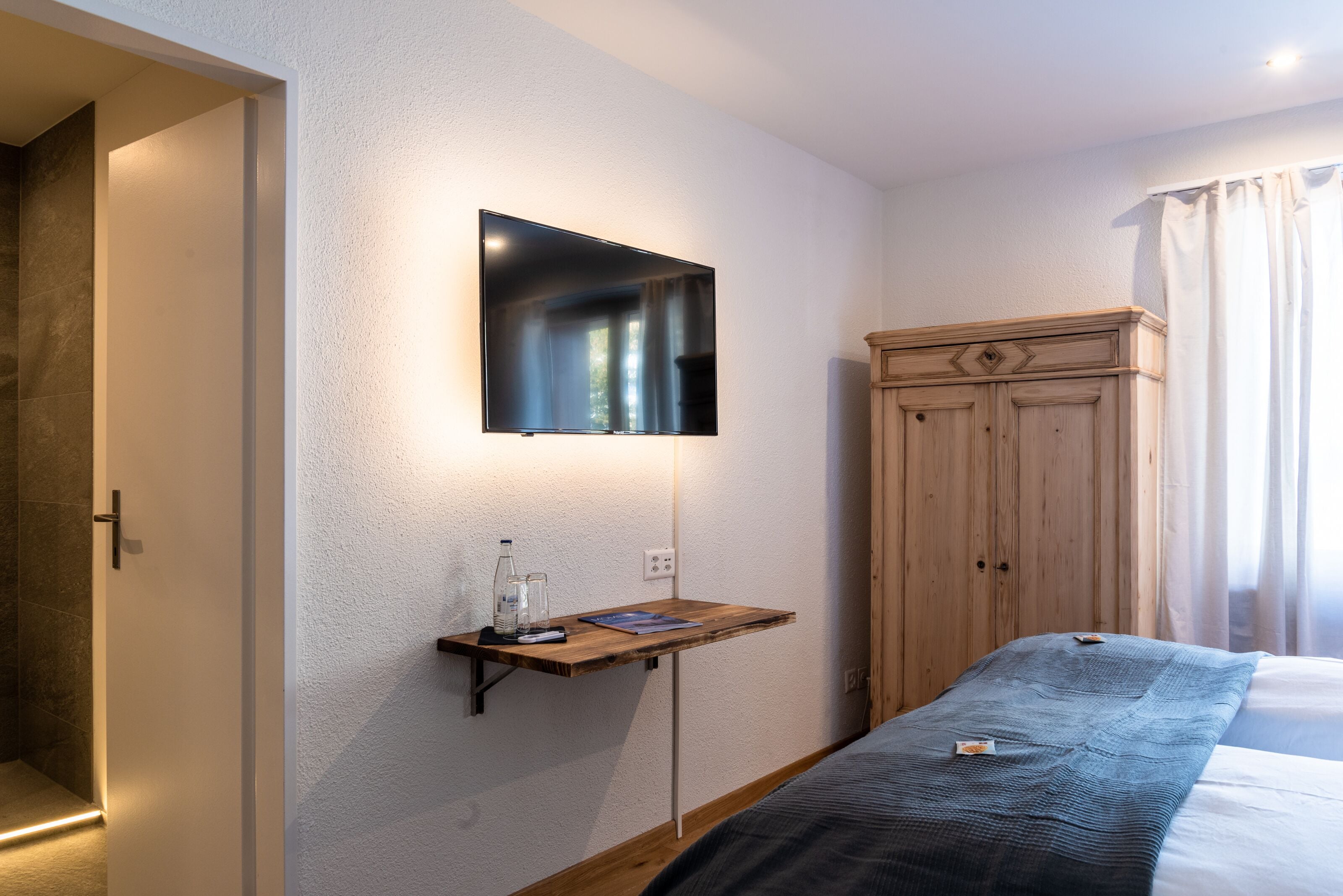 Habitación de hotel con cama, armario de madera, televisor montado y mesita de noche con una botella y vasos.