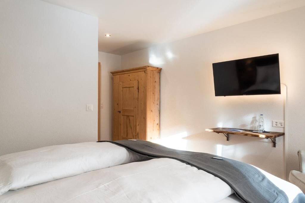 A bedroom with a bed, a wooden wardrobe, a mounted TV, and a shelf with bottles.