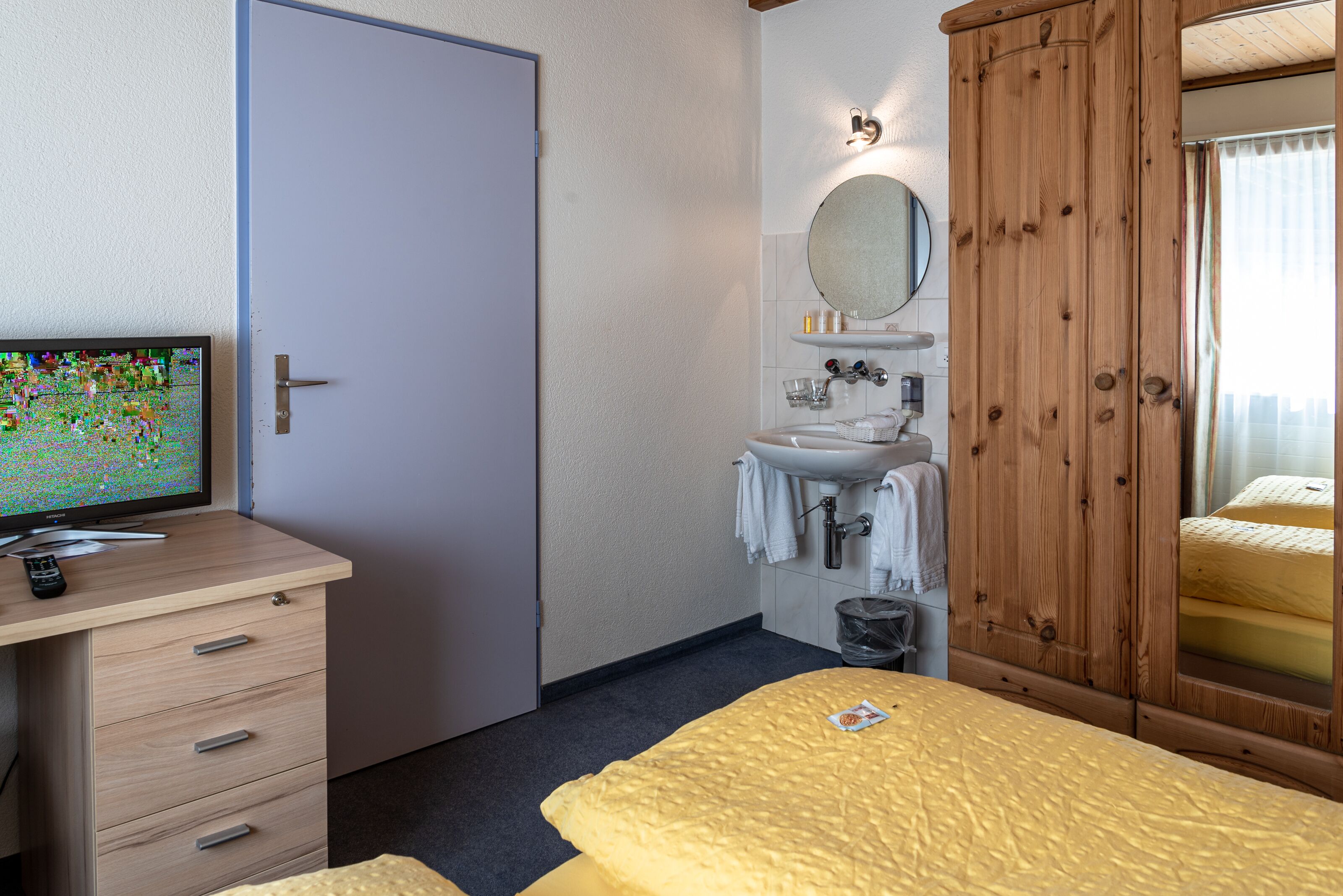 A hotel room with a bed, a wooden wardrobe, a bathroom with a sink and mirror, and a desk with a television.